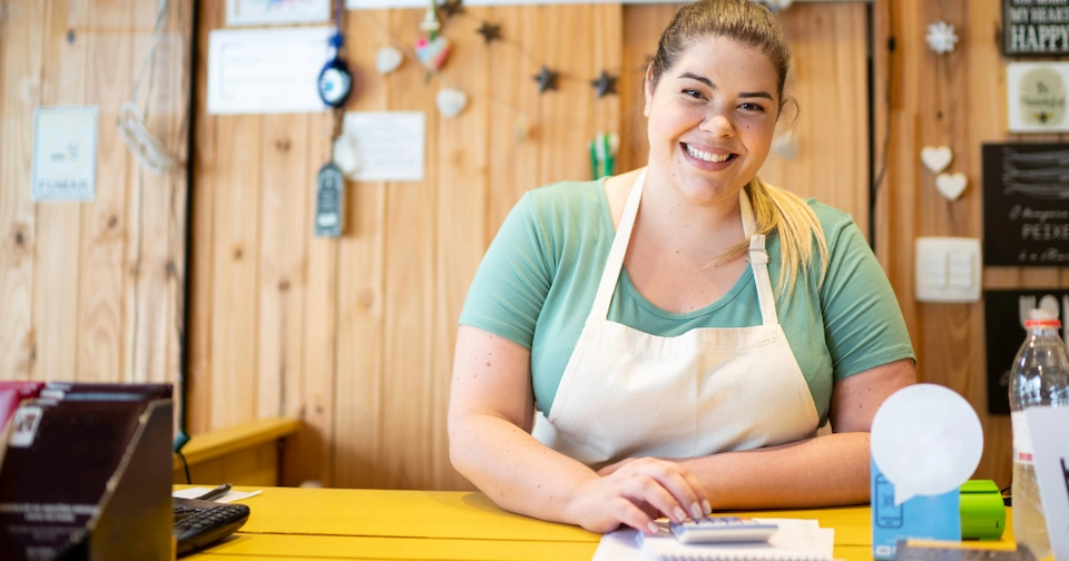 female small business owner smiling, wearing an apron, and working behind the counter